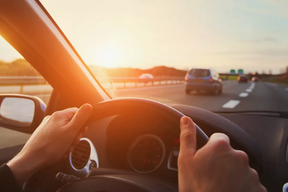 Cose-up of person's hands on steering wheel driving down highway with sunset sky.